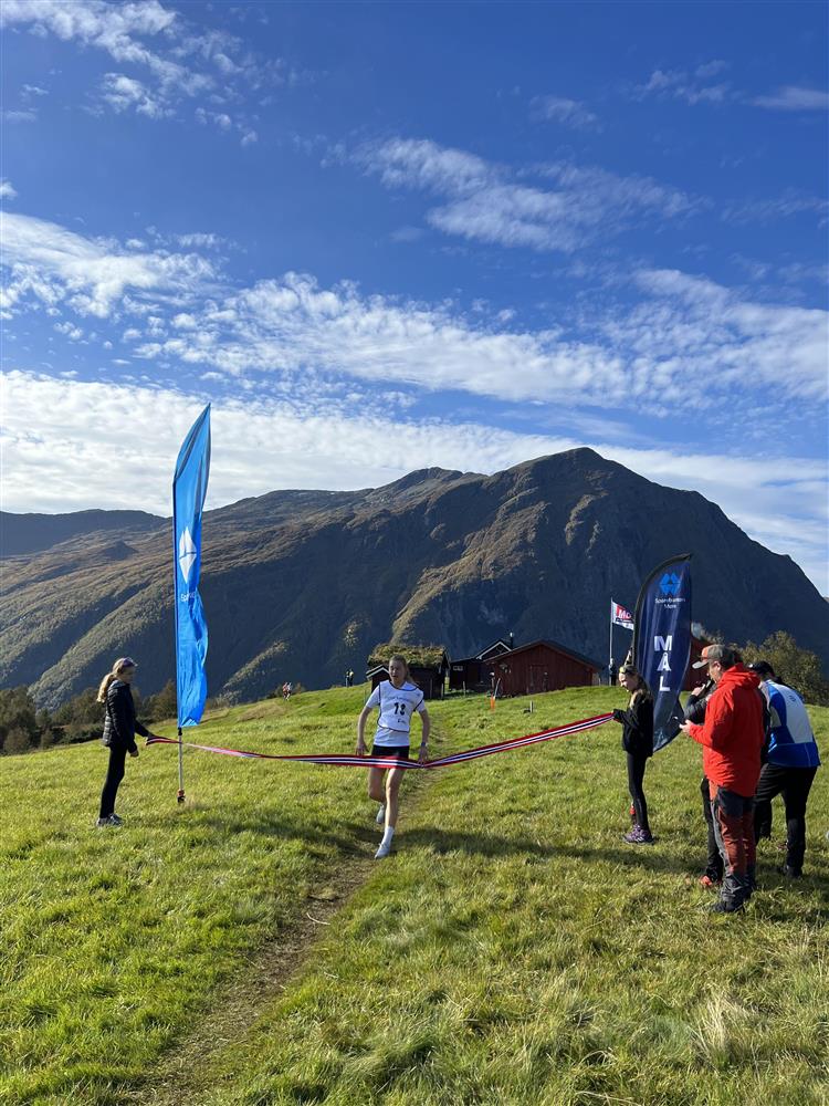 Ein deltakar passerer målseglet på ein setervoll i eit fjellområde. Nokre tilskodarar til stades. Blå himmel og sol. - Klikk for stort bilete