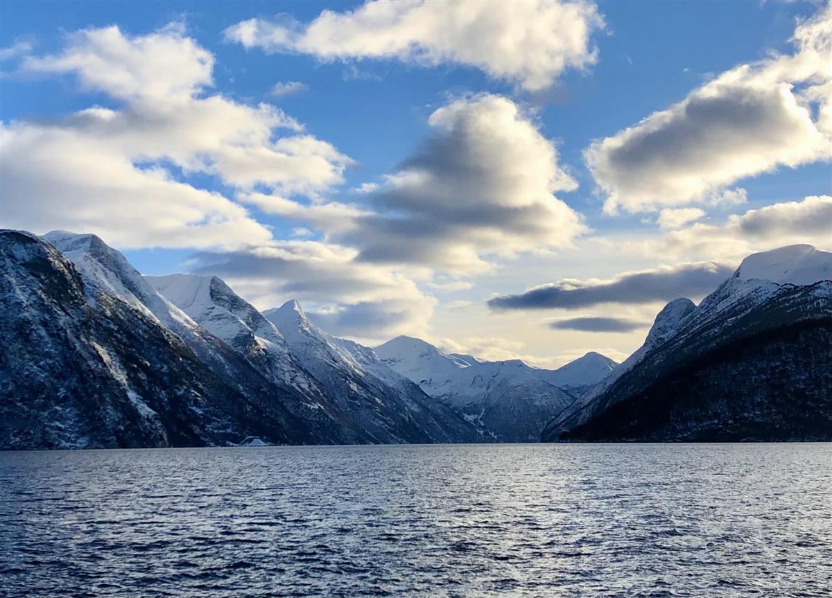 Ein trong fjord omkransa av bratte, snødekte fjell. Blå himmel med skyer. - Klikk for stort bilete