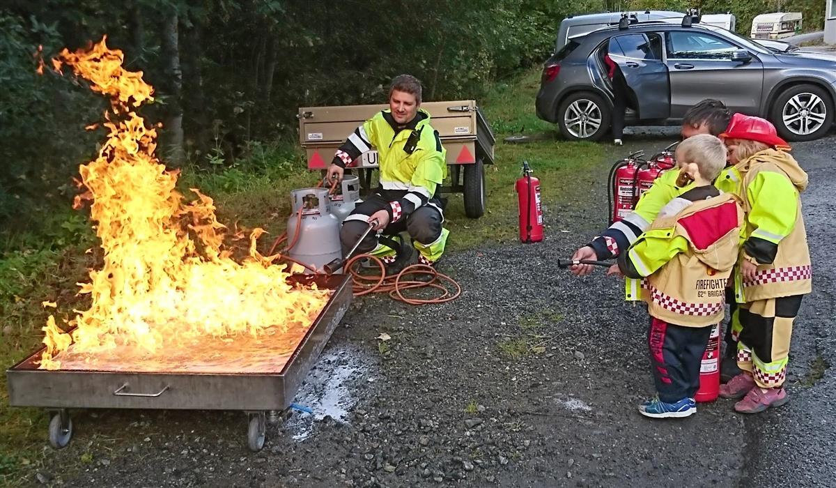 utandørs, brann i metallkasse, brannslokking, pulverapparat, person kledd i brannutstyr, barn som ser på 