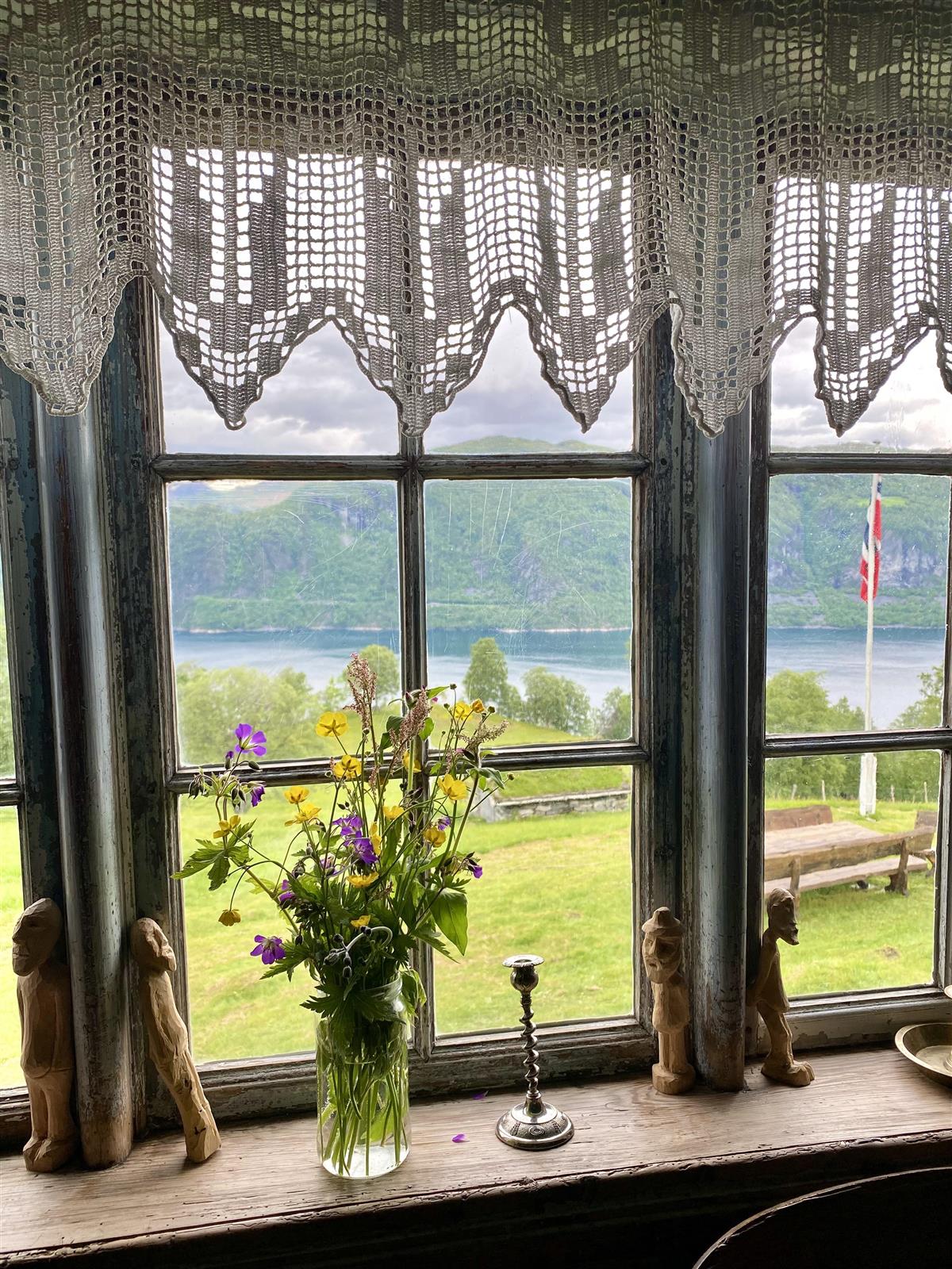 Eit gammalt vindu pynta med markblomster og trefigurar. Ute er flagget heist og det er utsikt til fjord og fjell. - Klikk for stort bilete