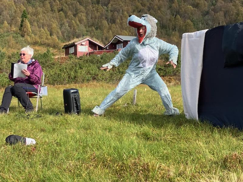 Ein person sit på ein campingstol med mikrofon og papir i hendene. Ein høgtalar står ved sidan av. Ei raud hytte i bakgrunnen. Grøn grasmark med haustfarga lauvskog i bakgrunnen.. - Klikk for stort bilete