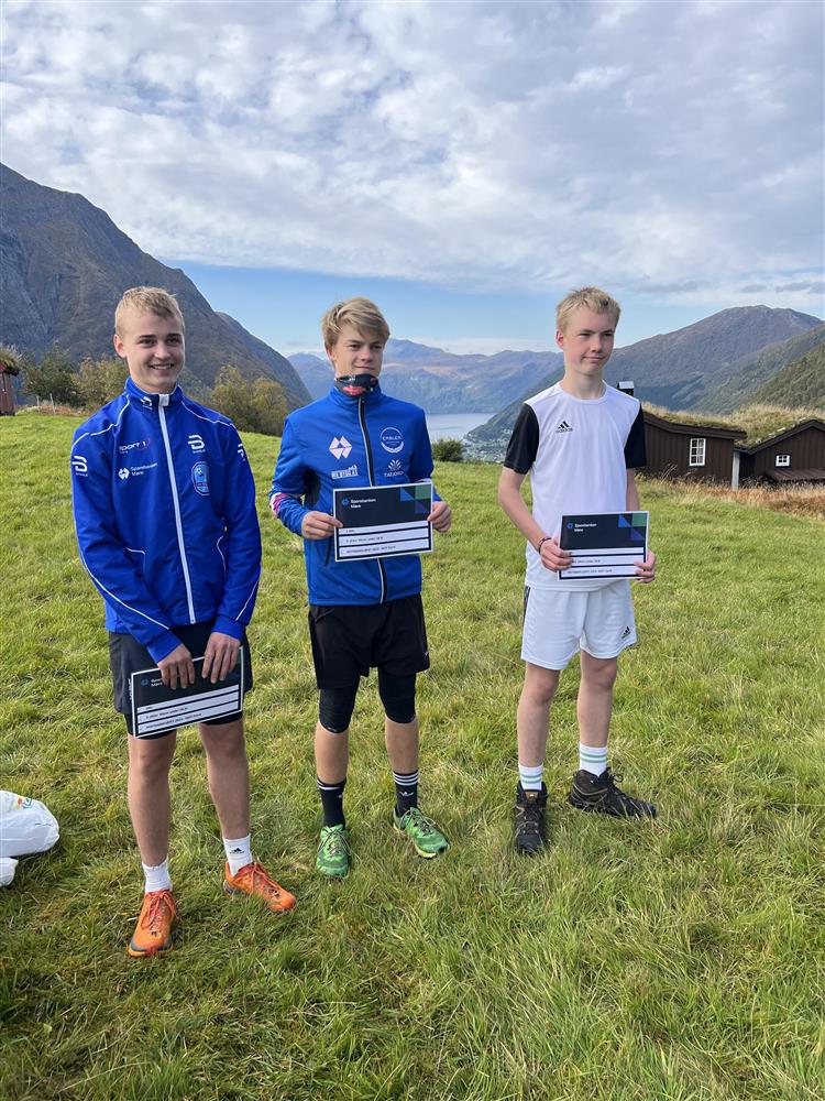 Tre treningskledde gutar med gåvesjekkar på ein setervoll i eit fjellområde. Blå himmel. - Klikk for stort bilete