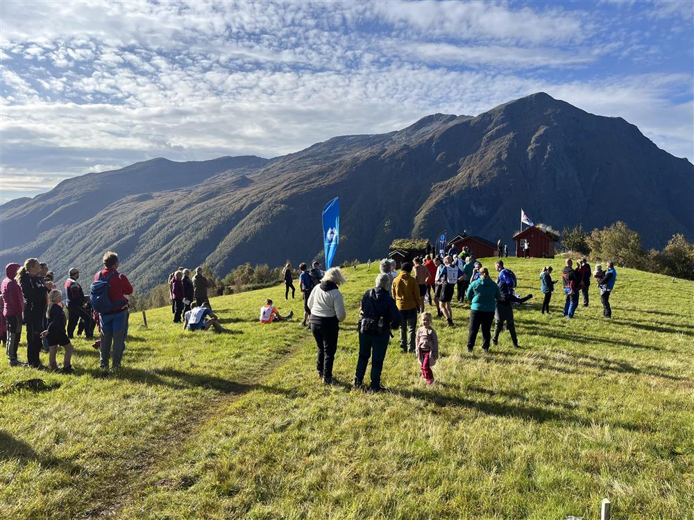 Mange personar samla rundt eit målsegl p ein setervoll i eit fjellområde. Blåh immel og sol. - Klikk for stort bilete